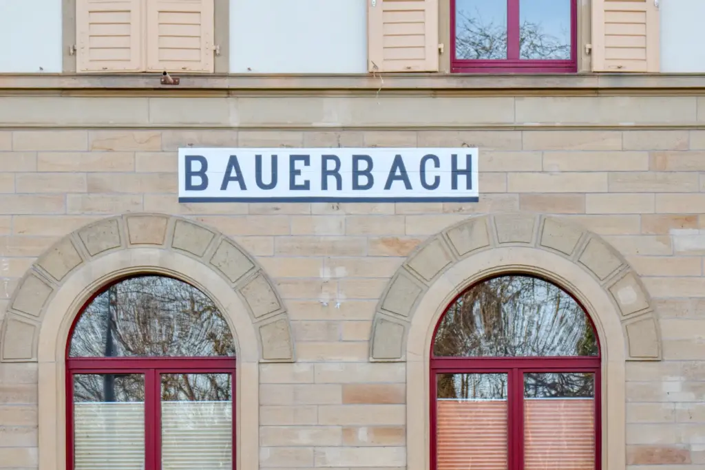 An einer Gebäudewand aus glatten Sandstein sieht man zwei Fenster mit roten Rahmen. Darüber steht Bauerbach.