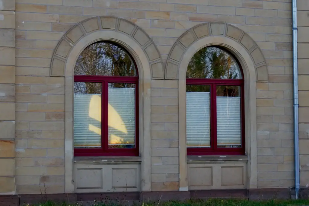 Wand eines Gebäudes aus glatten Sandstein mit zwei Fenster. Die Rahmen der Fenster sind rot gestrichen.