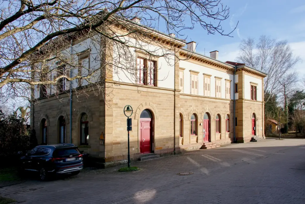 Seitliche Ansicht eines Gebäudes, was teilwiese durch unbelaubte Äste verdeckt wird. Das Obergeschoß ist weiß und das Erdgeschoß ist aus glatten Sandstein. Die Rahmen der Fenster sind rot. Links vom Gebäude steht ein Auto.