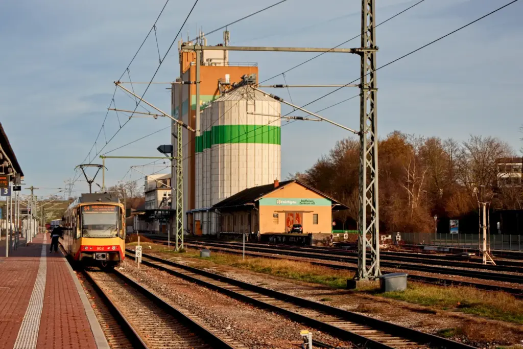 links steht eine Bahn an einem Bahsteig. Im Hintergrund sind riesige runde und eckige Silotürme für Getreide zu sehen.